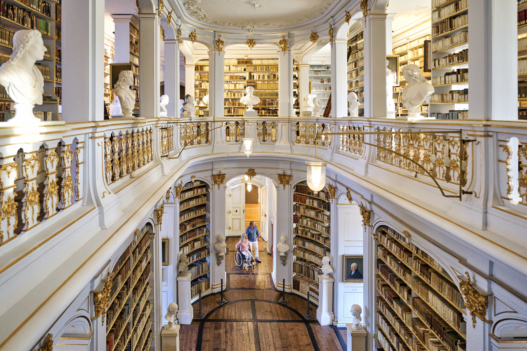 Rokokosaaal der Herzogin Anna Amalia Bibliothek Der barocke Rokokosaal ist zweistöckig und mit Weiß und Gold verziert. Mehrere Bücherwände stehen sich symmetrisch gegenüber und sind gefüllt mit einer Vielzahl an dunkelbraunen Lederbänden. Auf beiden Etagen blicken sich etwas mittig des Raumes gegenüberstehende weiße Büsten von bedeutenden Literaten an.