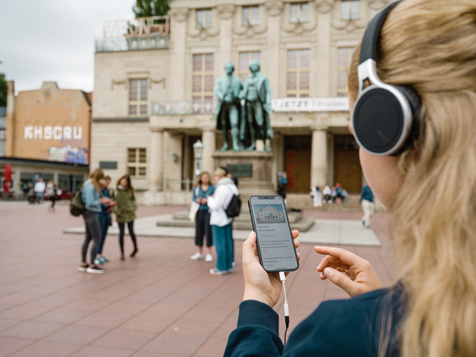 App Weimar+ Frau lauscht über die Kopfhörer einer Hörgeschichte zum Deutschen Nationaltheater, ihr Blick geht Richtung Goethe- und Schiller-Denkmal