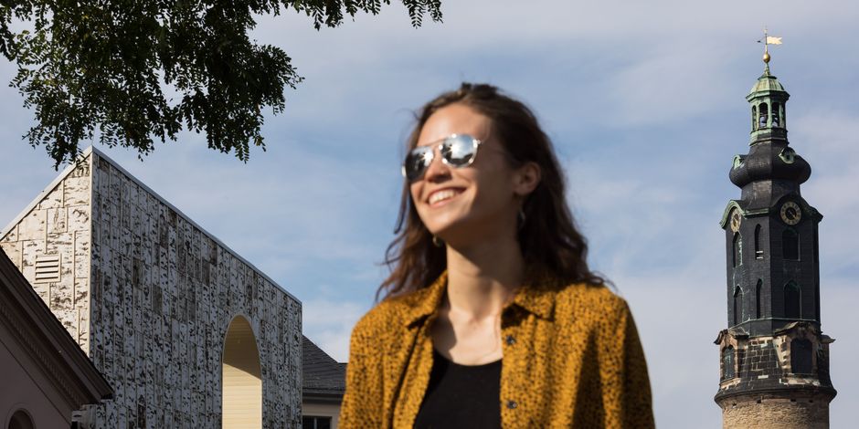 In the foreground there is a woman in sunglasses radiating joy and in the background there is a castle tower and a new building made of multi-layered solid wood panels