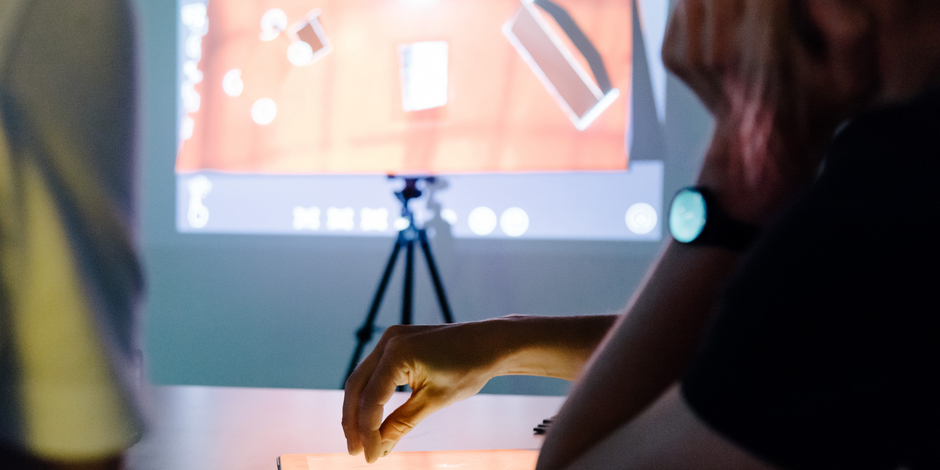 Dokumentation zum Video-Mapping Workshop Zwei Menschen bearbeiten etwas an einem Tablet. Im Hintergrund ist eine Projektion an die Wand zu sehen.