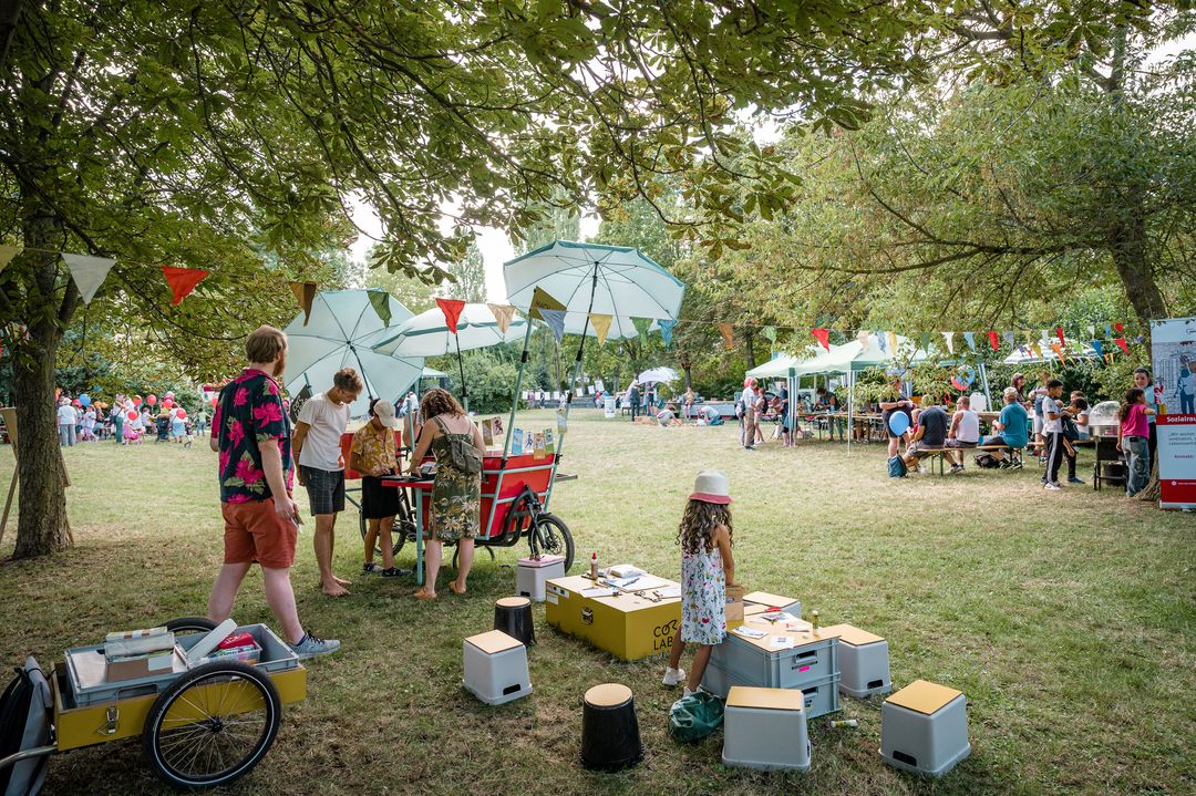 Co-Labor unterwegs Auf einer sommerlichen Wiese sind mehrere Zelte aufgebaut. Daneben steht ein Lastenrat mit Sonnenschirmen und Spielkisten. Dies ist das Co-Labor unterwegs.