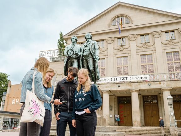 App Weimar+ Vor dem Goethe- und Schiller-Denkmal steht eine Gruppe von vier Personen, die gebannt auf ein Smartphone schauen und Inhalte der Kulturapp abspielen.