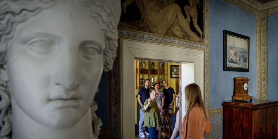 Riesige Büste von der Göttin Juno im Vordergrund, im Hintergrund Gäste, die sich durch Wohnräume bewegen