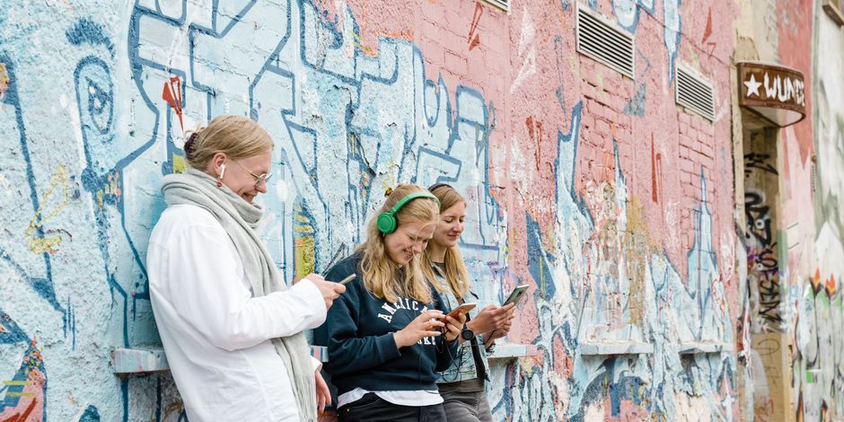 App Weimar+ A group of four people, standing in front of a graffiti-covered wall, listen and watch attentively to the content of the cultural app on a smartphone