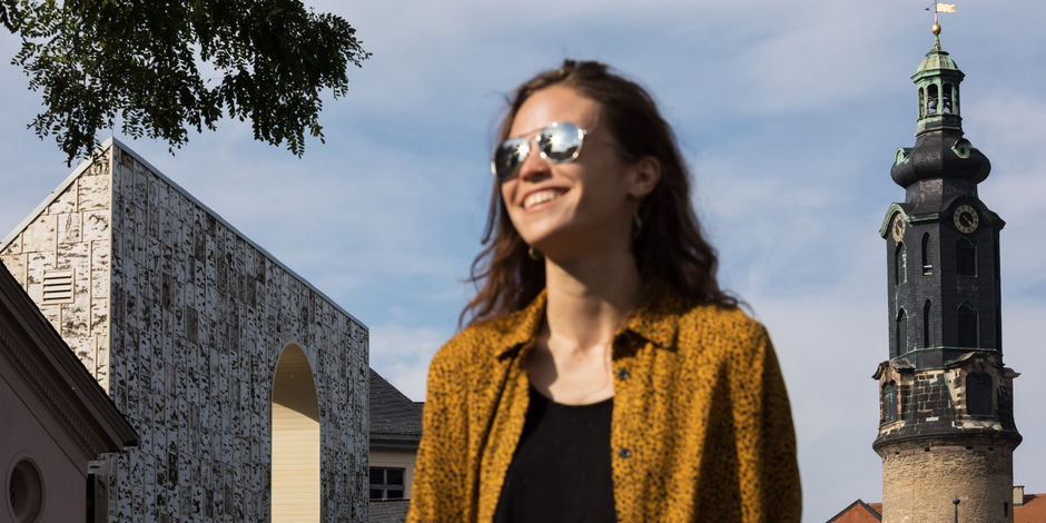 Im Vordergrund steht eine Frau mit Sonnenbrille, die Freude ausstrahlt und im Hintergrund befinden sich ein Schlossturm und einem Neubau aus mehrschichtigen Massivholzplatten